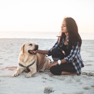 A woman sitting on the beach petting a happy golden retriever.